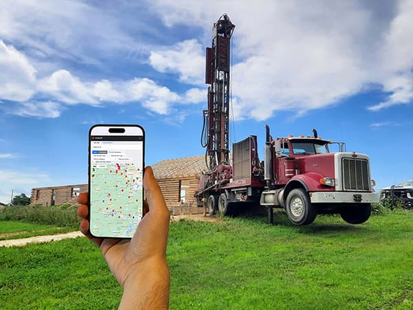 A driller holding a phone running the DrillerDB field app in front of the drilling rig